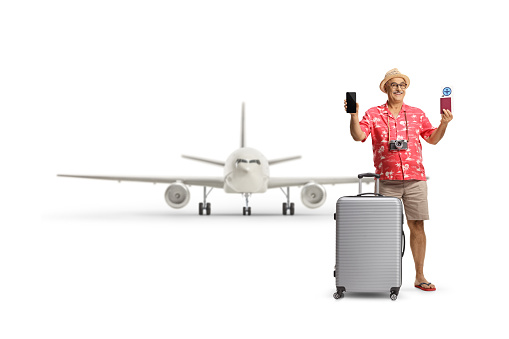 Mature male tourist with a suitcase showing a passport and a smartphone in front of an airplane isolated on white background