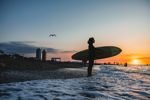 A female surfer in a wetsuit standing on a beautiful beach in Barcelona and holding her surfboard. She is admiring the beautiful view of the waves during a sunset. The sun is setting and the sky is orange.