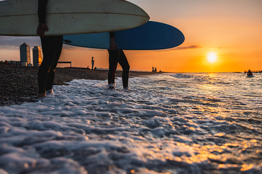 Close-up of two female surfers stepping into the water before a morning surf session during sunrise. Only legs and surfboards are visible, no face. The sun is rising and the sky is orange. Idyllic view of the beach and horizon. Beautiful surfing conditions.