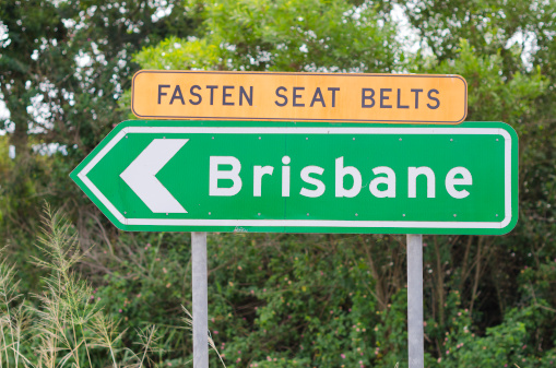 Green Brisbane sign with arrow on two posts. Above the green sign there is a yellow fasten seat belts sign. Background has out of focus bush.