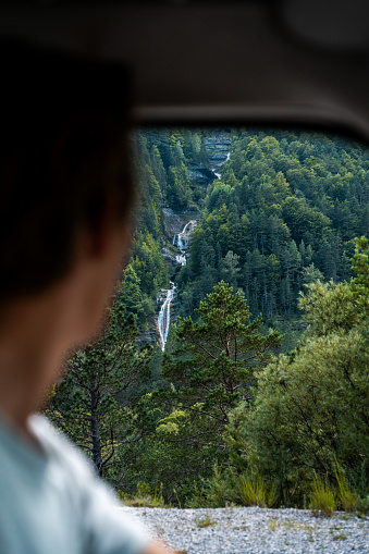 A man looking out of the window of his van at the mountainous landscape of a falling waterfall.