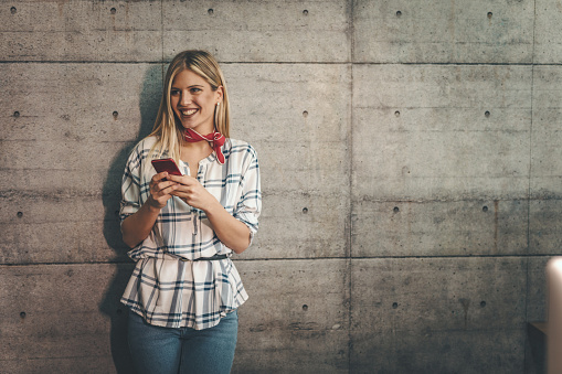 Young beautiful successful smiling female entrepreneuris writing messages on a smartphone in front of a concrete wall.