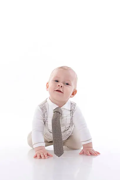 Smiling baby in a beige romper suit and tie crawling. On a white background with reflection