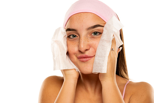 young happy woman with headband cleans her face with wet wipes on white background.