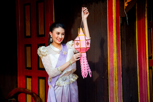 Young Asian woman with contemporary Thai dress stand and hold pink traditional lantern in area of ancient building and she look happy.