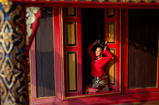 Side view of beautiful young Asian woman sit on red door and touch her hair look like to set hair style in area of ancient building of Northern Thailand with happiness.