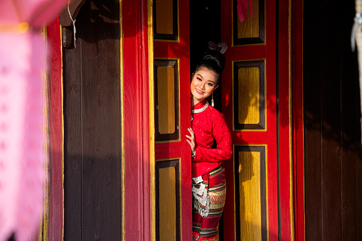 Front view of young Asian woman wear Thai Lanna dress stand in red color and old style door also look at camera with smiling in the area of ancient building.