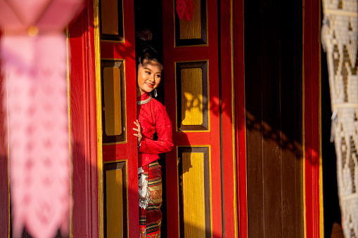 Young Asian woman wear Thai Lanna dress stand in red color and old style door also look to right side with smiling in the area of ancient building.