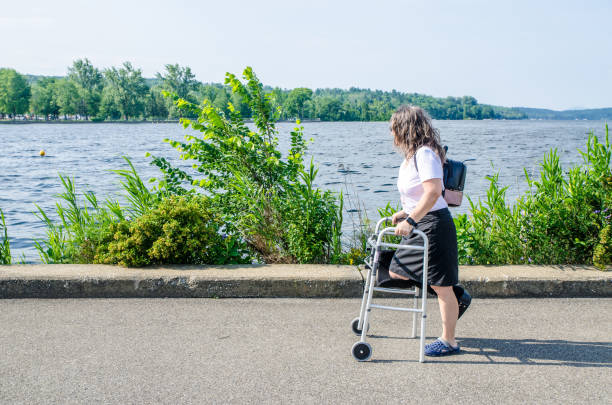 Disabled woman walking with walker and orthopedic boot besides Memphremagog lake during summer day