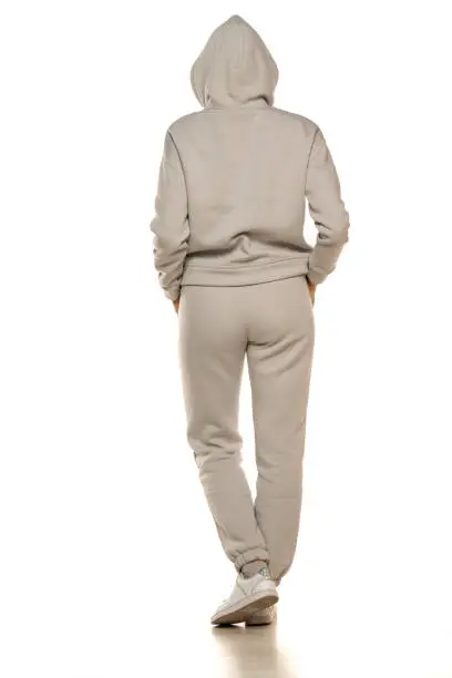 Photo of Rear view of a young woman in a gray tracksuit and hood posing to a white background in the studio