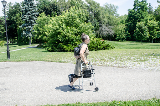 Disabled woman walking with walker and orthopedic boot in public park during summer day