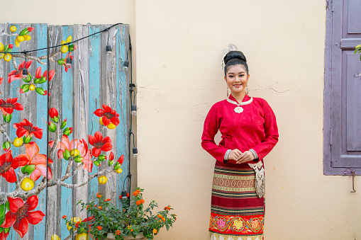 Portrait of young Asian woman with Thai lanna traditional dress stand near wall of ancient house that one of lanmark for tourist and she smile and look at camera.