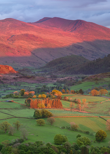 Beautiful evening light illuminating green countryside pastures with glowing red sunset light on Helvellyn mountain range in the Lake District, UK.