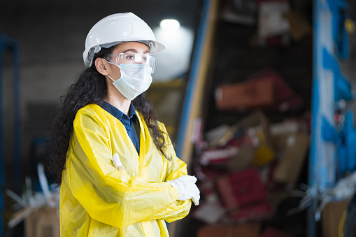 Black-haired female inspector standing against conveyor belt at waste recycling plant. Woman supervisor managing waste sorting process