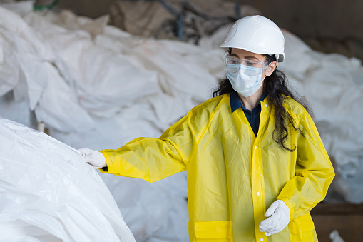 Black-haired lady worker in mask and protecting glasses sorts trash in waste processing plant. Supervisor prepares garbage for sorting in factory yard