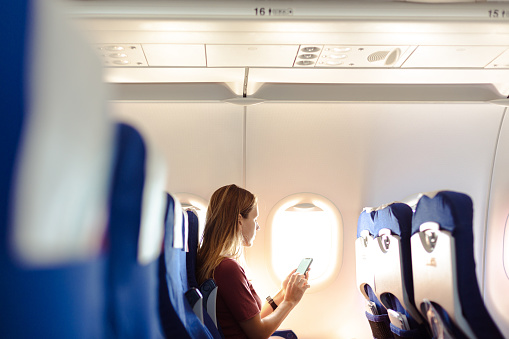 Young woman sitting on the aircraft seat near the window during the flight in the airplane. She is using smart phone with a blank screen