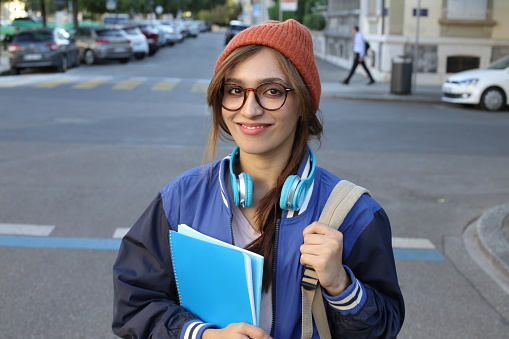 A very beautiful young female student in her twenties is walking on the street with her backpack. She wears an orange beanie and a classic university jacket.