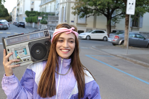 A gorgeous South Asian woman in her twenties is listening to music on the street. She uses a vintage stereo cassette and wears a colorful outfit.