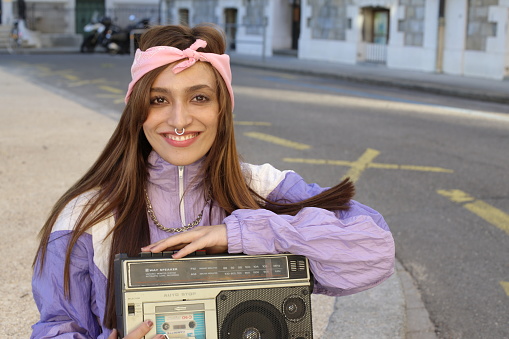 A gorgeous South Asian woman in her twenties is listening to music on the street. She uses a vintage stereo cassette and wears a colorful outfit.