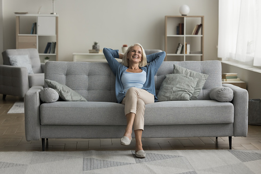 Happy elderly woman relaxing on grey soft couch at cozy home, breathing fresh air, looking at window away, thinking, daydreaming, smiling, laughing, enjoying comfort, leisure in modern interior