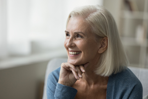 Cheerful positive blonde senior woman sitting on sofa, thinking on good retirement plans, touching chin, looking away, smiling, laughing. Happy elderly lady enjoying relaxation, leisure at home