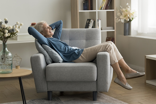 Happy joyful senior woman resting in soft cozy armchair at home, smiling, laughing with closed eyes, enjoying comfort, leisure, weekend in contemporary interior, breathing fresh cool air