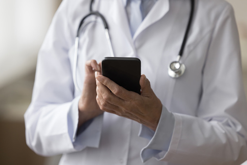 Cropped shot of doctor woman in uniform coat using medial application on smartphone for online connection, giving consultation to patient, holding mobile phone, touching screen, texting, typing
