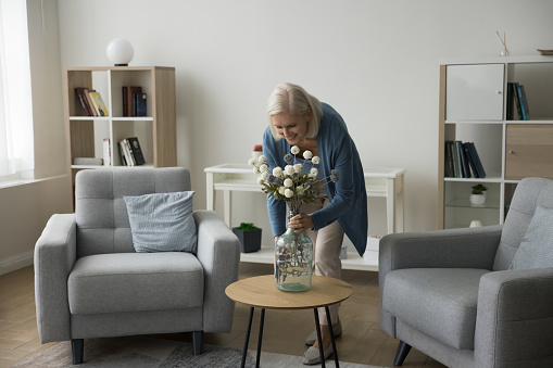 Happy blonde retired mature woman decorating home living room interior, placing flowers in glass bottle vase, smiling, enjoying care of apartment design, floristic hobby