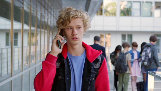 Schoolboy walking on street to school and talking on cellphone. Portrait of caucasian teenage boy student having phone call walking outdoors. High quality photo