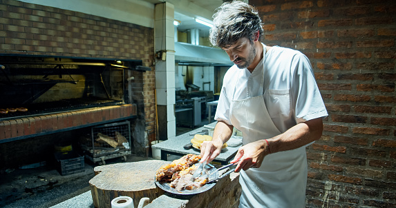 A handsome chef in his 40s cutting Argentinian meat with a knife and a tong in the kitchen of a restaurant.