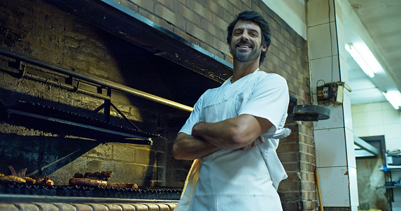 A handsome chef in his 40s looking at the camera while posing in front of a grill in the kitchen of a restaurant.