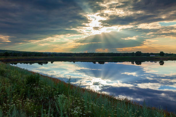 A cloudy sky with the rays of the sun breaking through the clouds and falling on a meadow covered with wild flowers, a pond and buildings visible in the distance. The wetlands near small town Rubizhne, Lugansk region, Ukraine.