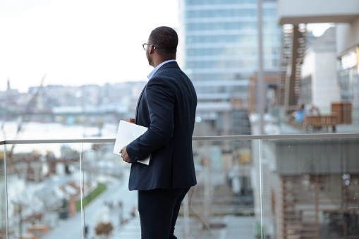 A young African-American businessman on the balcony holding a laptop and looking straight