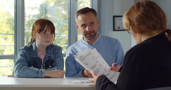 Back view of female headmaster interview father and preteen girl in school office. Smiling mature man with teenage daughter meeting woman principal