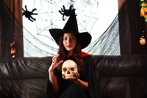 beautiful young woman in a witch Halloween costume wears a witches hat holding a human skull over the spooky dark magic background - Halloween party concept.