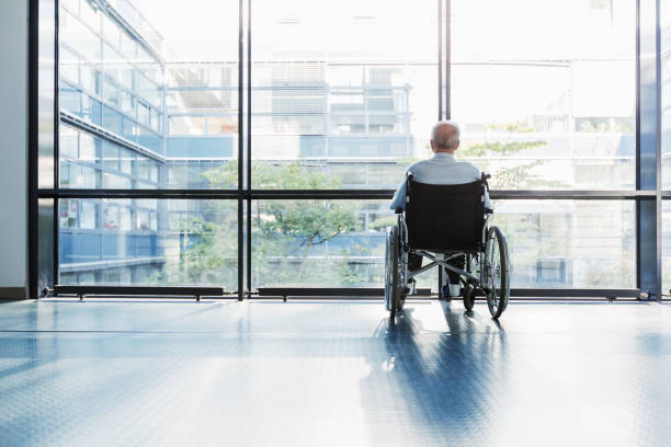 Senior Man in Wheelchair Senior Man in Wheelchair looking out of a window in a hospital corridor. medical supplies wheelchair medical equipment nursing home stock pictures, royalty-free photos & images
