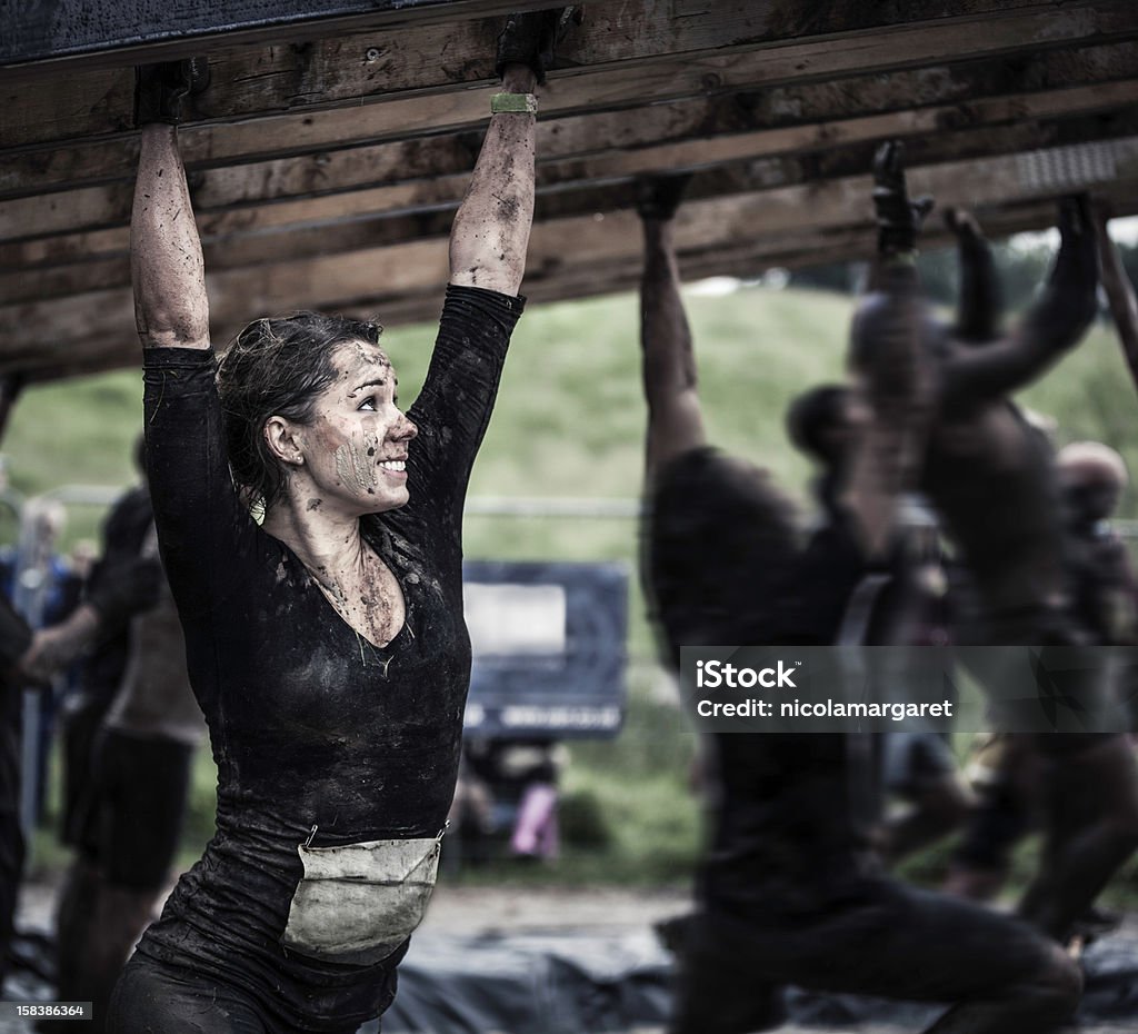 Female athlete competing in an obstacle course Young woman on monkey bars covered in mud. Obstacle Course Stock Photo