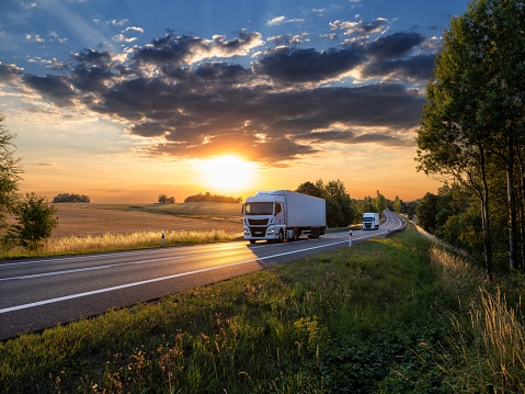 Two white trucks driving on the asphalt road in rural landscape with dramatic cloud at sunset