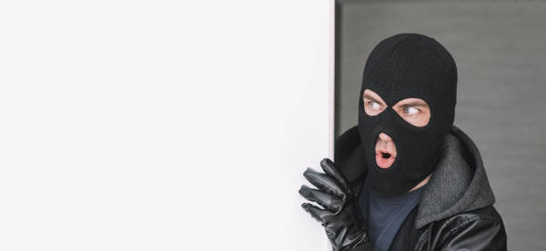 istock An evil thief in mask looks out from behind a white board. Space for text and design. copy space. the surprised bandit looks away 1583957084