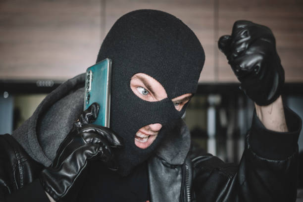 thief is talking on phone in black mask. the extortionist demands money for the kidnapping. a phone fraudster commits a crime, violates the law. concept of telephone terrorism and fraud. phone threats - extortionist imagens e fotografias de stock