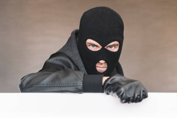 istock An evil thief in a mask looks out from behind a white board. evil bandit with a mask on his face. 1583957150