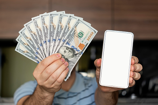 man wears casual clothes blue shirt in hand fan of cash money in dollar banknotes use mobile cell phone. an empty white screen on a smartphone to advertise the app.