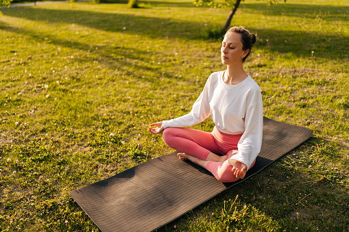 High-angle view of yogini female during relaxation and meditation in park meditation session. Chilled-out young woman with close eyes and concentrated face expression holding hands on knees.