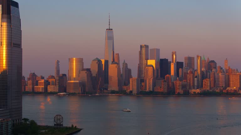 Lower Manhattan with One World Trade Center distant view over the Hudson River with boats sailing along the river, seen from Paulus Hook in Jersey City at sunset. Aerial video with the backward camera motion.