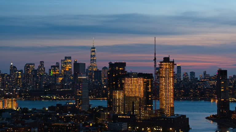 New building construction in Greenpoint, Brooklyn, illuminated at night, with Newtown Creec flowing into the East River. Distant view of Manhattan with visible Freedom Tower. Aerial video with the panning-descending camera motion.