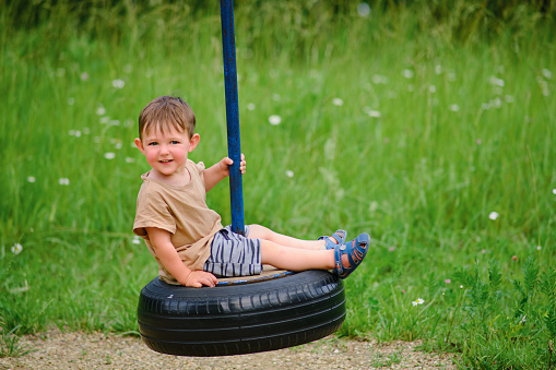 A smiling child enjoys playing on a car tire turned into a swing. A happy baby is spinning on a swing made of a tire from a car wheel. Kid aged two years