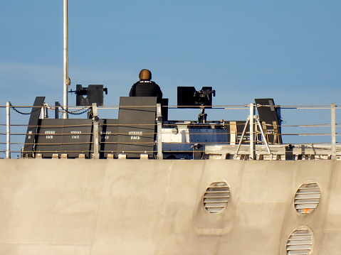 A manned machine gun post on the forward deck of USS Canberra (LCS 30), an Independence Class combat ship of the US Navy docked at Garden Island in Sydney Harbour.  She was in port for her commissioning ceremony on 22 July 2023. The flagpole is to display the Jack of the United States.  Two machine guns are surrounded by bulletproof protective plates stenciled with \