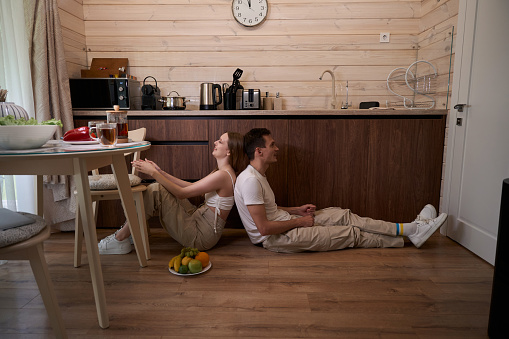 Man and a woman sit with their backs to each other on the floor in the kitchen