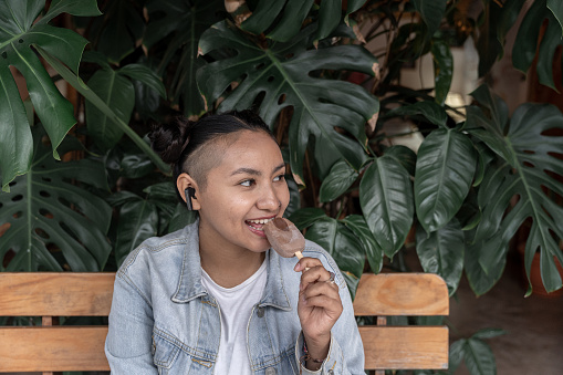 A young Hispanic woman is enjoying an ice cream while listening to music sitting on a bench with plants in the background. Concept of sweet snack and technology