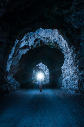 A lone man walks down a dark tunnel with a headlamp on